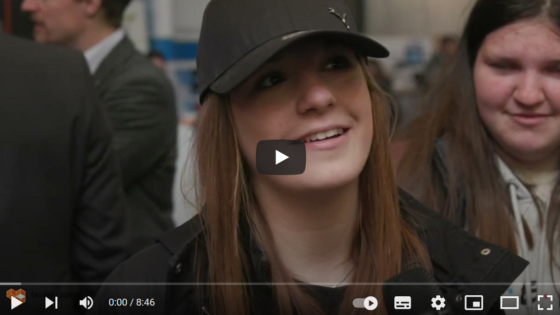 YouTube-Titelbild: Junge Frau mit langen braunen Haaren und schwarzer Baseballkappe lächelt, im Hintergrund weitere Personen in einer belebten Innenraumszene.