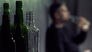 Mehrere leere Glasflaschen im Vordergrund; im unscharfen Hintergrund trinkt eine Person aus einer Flasche.