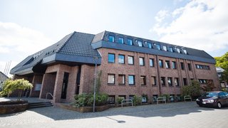 Das Bild zeigt das Rathaus von Niederkrüchten, ein großes Backsteingebäude mit dunklem, schrägem Ziegeldach und mehreren kleinen Dachgauben. Die Fassade besteht aus rotbraunen Klinkersteinen und zahlreichen rechteckigen Fenstern. Links führt eine kurze Treppe zum überdachten Eingangsbereich, rechts stehen einige Sträucher und ein geparktes Auto. Der Vorplatz ist mit Pflastersteinen ausgelegt, darüber ein heller Himmel mit vereinzelten Wolken.“