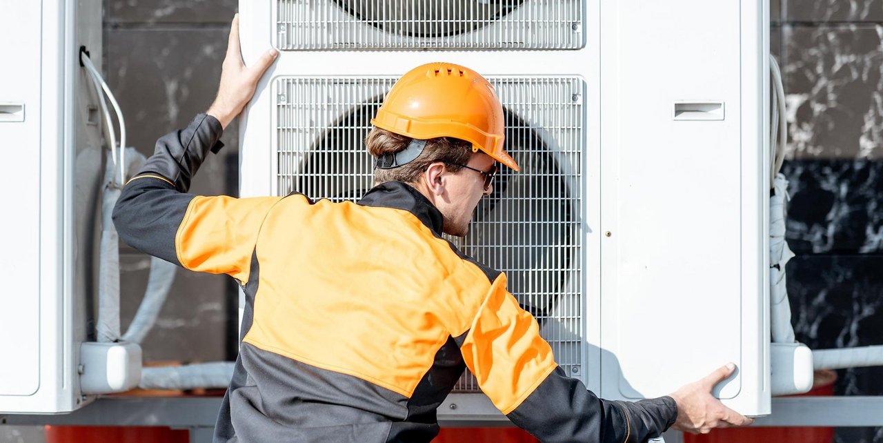 Ein Handwerker mit orangefarbenem Helm und Arbeitsjacke überprüft eine große Außenklimaanlage mit zwei Ventilatoren.