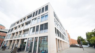 Das Bild zeigt das Stadthaus in Viersen. Das mehrgeschossige Verwaltungsgebäude hat eine helle Fassade mit großen rechteckigen Fenstern und einer modernen, klaren Architektur. Im Erdgeschoss befinden sich verglaste Eingänge und eine überdachte Arkade mit Säulen. Vor dem Gebäude stehen Fahrräder, daneben erstreckt sich ein gepflasterter Platz mit Treppen und Bäumen. Der Himmel ist bewölkt.