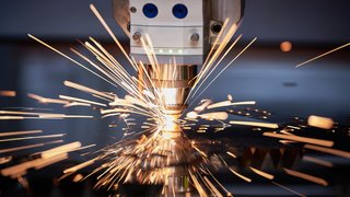 Nahaufnahme eines industriellen Laserschneidgeräts, das mit hoher Präzision Metall bearbeitet. Helle, orangefarbene Funken fliegen in alle Richtungen und erzeugen einen dynamischen Eindruck von Energie und Technik. Die Szene findet in einer modernen Fertigungsumgebung statt.