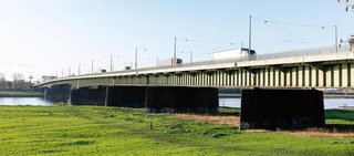 Die Josef-Kardinal-Frings-Brücke über den Rhein bei Tageslicht. Fahrzeuge, darunter Lkw, überqueren die Brücke. Im Vordergrund eine grüne Wiese mit einer spazierenden Person und einem Hund, klare Sicht auf Brückenpfeiler und stählerne Tragstruktur.