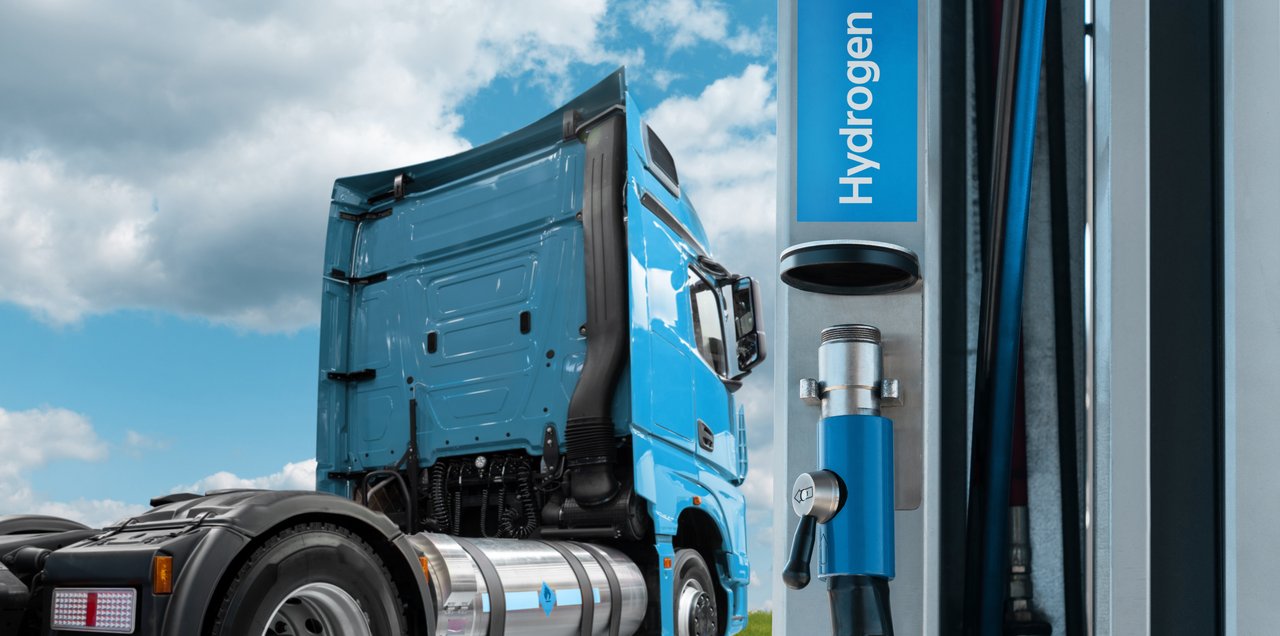 Blauer Lkw an einer Wasserstofftankstelle; im Vordergrund ist die Zapfsäule mit Wasserstoff-Anschluss sichtbar.