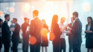 Silhouetten von Geschäftsleuten in formeller Kleidung, die in kleinen Gruppen bei einer Netzwerkveranstaltung oder einem Empfang in einem hell erleuchteten Raum miteinander sprechen. Die Szene ist unscharf und vermittelt eine lebendige, kommunikative Atmosphäre.