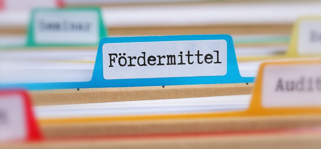 Reiter mit der Aufschrift „Fördermittel“ in einer Hängeregistratur, umgeben von weiteren farbigen Registerkarten mit unterschiedlichen Beschriftungen, unscharf im Hintergrund.
