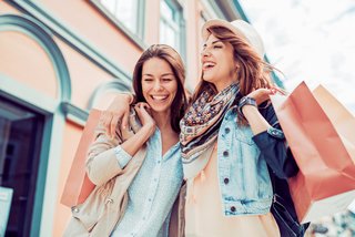 Zwei junge Frauen lachen und gehen Arm in Arm beim Einkaufen durch eine farbenfrohe Innenstadt. Eine trägt Einkaufstaschen, beide wirken fröhlich und entspannt.