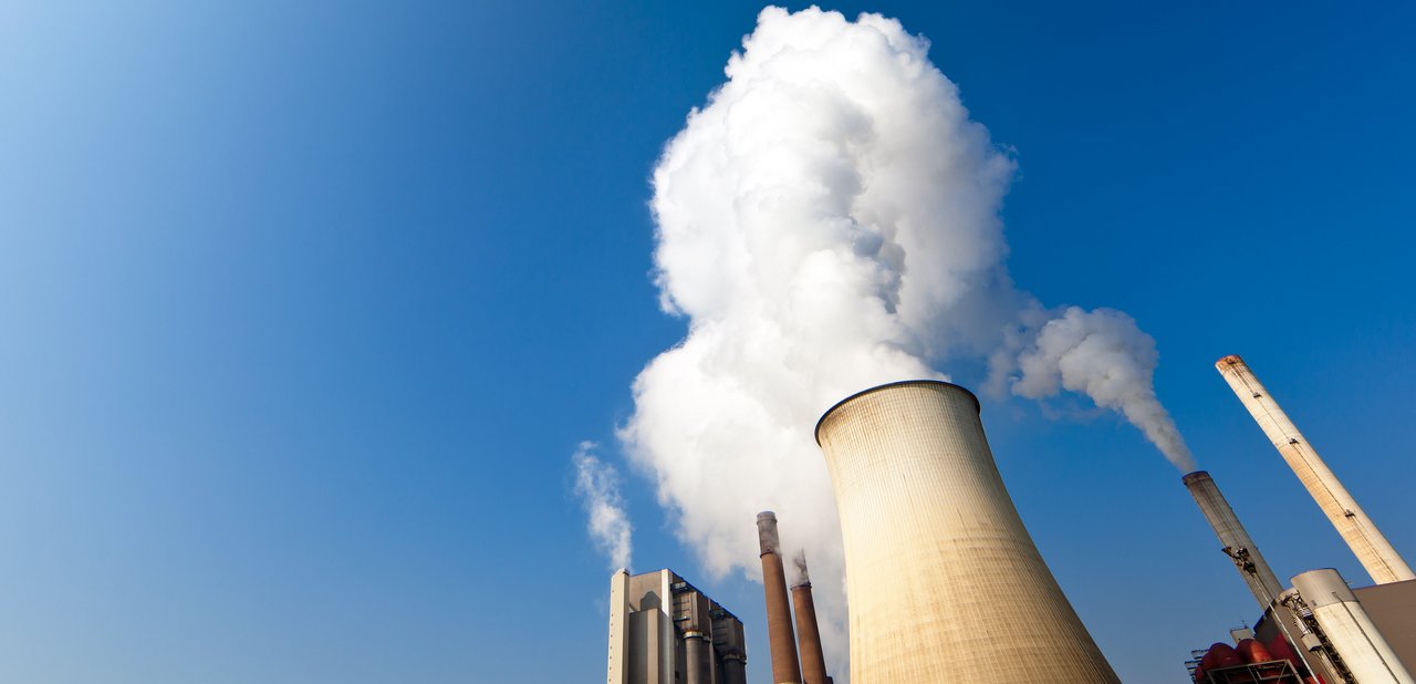 Das Bild zeigt ein großes Industriekraftwerk mit mehreren Schornsteinen, aus denen weißer Rauch oder Wasserdampf in den Himmel steigt. Besonders auffällig ist ein großer, konisch geformter Kühlturm, der eine dichte, weiße Dampfwolke ausstößt. Der Himmel ist klar und blau, was die weißen Dampfwolken besonders hervorhebt.