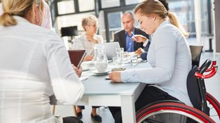 Eine Gruppe von Menschen in einem modernen Besprechungsraum. Im Vordergrund sitzt eine junge Frau im Rollstuhl an einem Konferenztisch und arbeitet konzentriert. Im Hintergrund sind Männer und Frauen unterschiedlichen Alters in ein Gespräch vertieft. Auf dem Tisch stehen Wassergläser, eine Karaffe und Arbeitsmaterialien. Die Szene zeigt ein inklusives und professionelles Arbeitsumfeld.