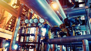 Das Bild zeigt eine industrielle Anlage im Inneren einer Fabrikhalle. Zahlreiche glänzende Rohrleitungen, Ventile, Druckanzeigen und Metallkonstruktionen durchziehen die Szenerie. Mehrere große Druckmesser sind in einer Reihe angeordnet, verbunden mit komplexer Technik. Warmes Licht von oben und seitlich erzeugt farbliche Kontraste aus Orange- und Blautönen und betont die industrielle Atmosphäre.