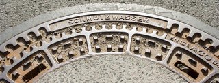 Teil eines runden Kanaldeckels aus Metall auf Asphalt, mit der erhabenen Aufschrift "SCHMUTZWASSER".