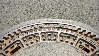 Teil eines runden Kanaldeckels aus Metall auf Asphalt, mit der erhabenen Aufschrift "SCHMUTZWASSER".