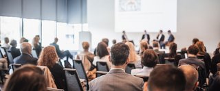 Teilnehmende sitzen in einem modernen Konferenzraum und verfolgen eine Podiumsdiskussion mit vier Personen auf einer Bühne im Hintergrund. Die Veranstaltung findet in heller Umgebung mit Tageslicht statt, Fokus liegt auf den Zuhörer:innen im Vordergrund.