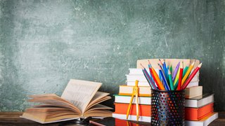 Ein hölzerner Schultisch vor einer grünen Tafel. Auf dem Tisch liegen ein aufgeschlagenes Buch, ein geschlossenes Smartphone, eine Lupe und ein Stapel Bücher mit bunten Einbänden. Daneben steht ein schwarzer Stiftehalter mit vielen farbigen Buntstiften. Ein gelber Zirkel lehnt an den Büchern.