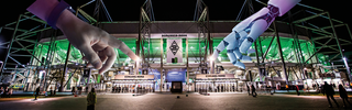 Fotomontage des Borussia-Parks bei Nacht. Vor dem Stadion zeigen eine menschliche Hand und eine Roboterhand aufeinander und symbolisieren die Zusammenarbeit von Mensch und Künstlicher Intelligenz.