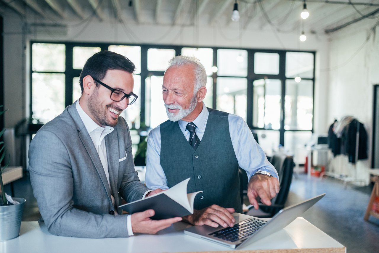 Zwei Männer in Businesskleidung stehen in einem modernen Büro. Der ältere Mann mit grauem Haar, Bart und Weste zeigt auf den Bildschirm eines Laptops, während der jüngere Mann mit Brille und Anzug ein Notizbuch hält und lächelt. Beide wirken in ein freundliches Gespräch vertieft.