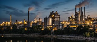 Große Industrieanlage bei Abenddämmerung, beleuchtete Gebäude und Schornsteine stoßen Rauch aus, Spiegelung im ruhigen Fluss im Vordergrund.