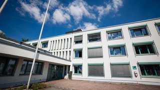 „Außenansicht des Rathauses in Jüchen, ein modernes weißes Verwaltungsgebäude mit mehreren Fensterreihen und einem offenen Vorplatz unter blauem Himmel.“