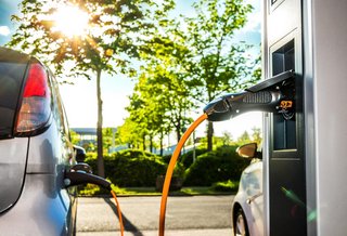 Ein silberfarbenes Elektroauto wird an einer öffentlichen Ladestation aufgeladen. Ein orangefarbenes Ladekabel verbindet das Fahrzeug mit der Station. Im Hintergrund sind grüne Bäume, Büsche und Sonnenschein zu sehen, was eine umweltfreundliche und moderne Mobilitätsszene vermittelt.