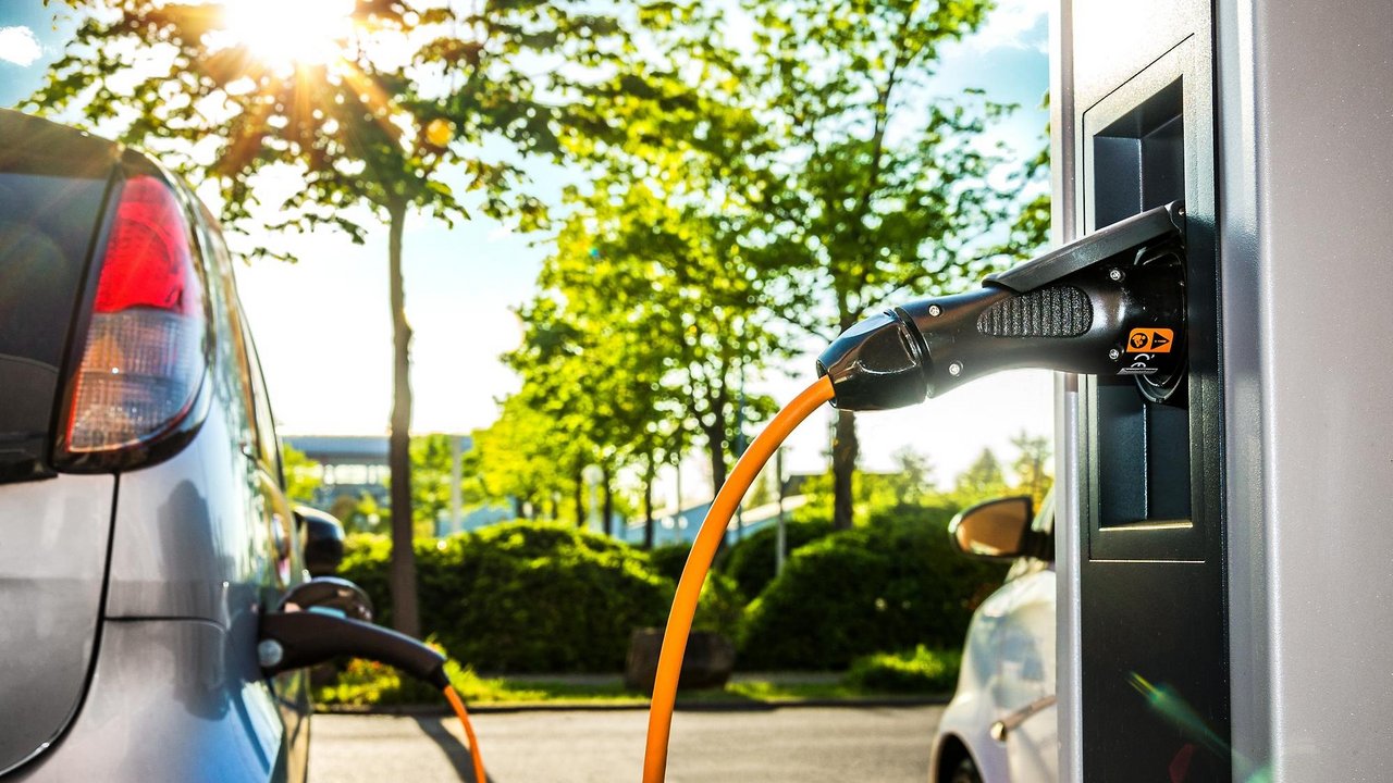 Ein silberfarbenes Elektroauto wird an einer öffentlichen Ladestation aufgeladen. Ein orangefarbenes Ladekabel verbindet das Fahrzeug mit der Station. Im Hintergrund sind grüne Bäume, Büsche und Sonnenschein zu sehen, was eine umweltfreundliche und moderne Mobilitätsszene vermittelt.