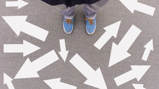 Person steht auf Asphaltboden, umgeben von zahlreichen weißen Pfeilen, die in verschiedene Richtungen zeigen – symbolisiert Entscheidungsfindung oder Orientierungslosigkeit.