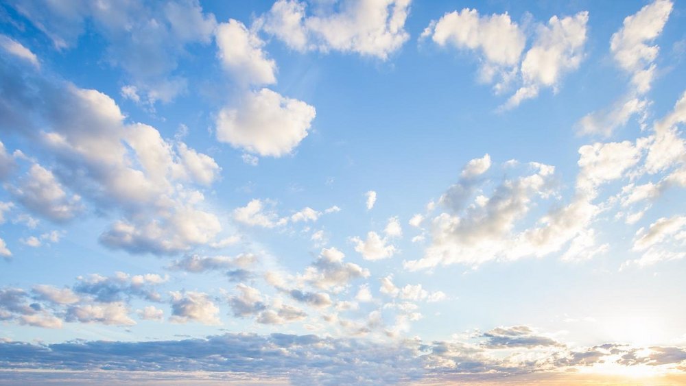 Weitwinkelaufnahme eines hellblauen Himmels mit zahlreichen, locker verteilten weißen und grauen Wolken bei Sonnenaufgang oder Sonnenuntergang, die Sonne ist tief am Horizont sichtbar und von einem sanften Lichtschein umgeben.