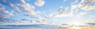 Weitwinkelaufnahme eines hellblauen Himmels mit zahlreichen, locker verteilten weißen und grauen Wolken bei Sonnenaufgang oder Sonnenuntergang, die Sonne ist tief am Horizont sichtbar und von einem sanften Lichtschein umgeben.