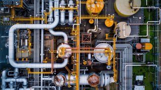 Drohnenaufnahme einer Industrieanlage mit zahlreichen Rohren, Tanks und metallischen Strukturen. Gelbe Wartungsstege und Rohrleitungen verlaufen über die gesamte Anlage, die aus verschiedenen runden und rechteckigen Einheiten besteht.