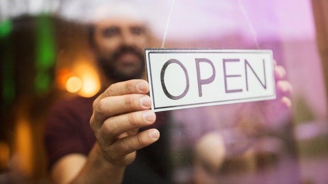 Ein Mann hängt hinter einer Glasscheibe ein Schild mit der Aufschrift „OPEN“ auf, das signalisiert, dass das Geschäft geöffnet ist. Die Szene ist unscharf im Hintergrund, der Fokus liegt auf dem Schild und der Hand des Mannes.