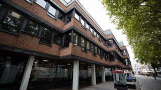 Backstein-Gebäude mit versetzten Fensterreihen und dunklem Fensterrahmen; im Vordergrund Glasvitrine mit der Aufschrift „Bridge-Club Kempen“, gepflasterter Gehweg mit Bäumen und Sitzbänken, Menschen im Hintergrund.