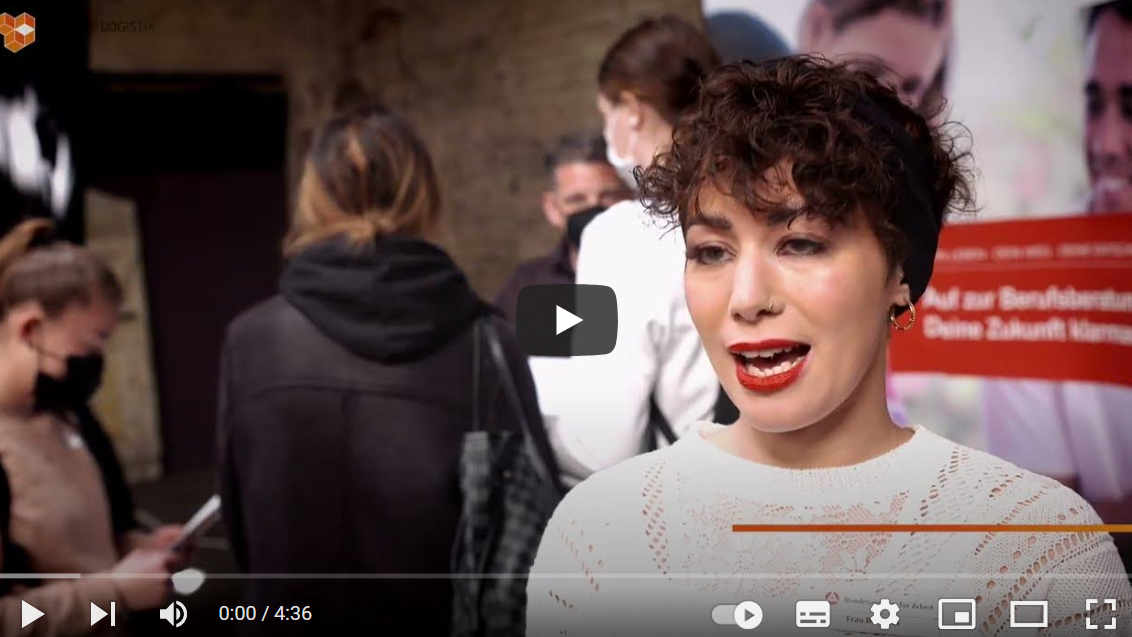 YouTube-Titelbild: Eine Frau mit kurzen, lockigen Haaren und rotem Lippenstift spricht in die Kamera. Im Hintergrund stehen mehrere Personen, einige tragen Masken. Links ist das Logo von „Logistik“ zu sehen, rechts ein rotes Banner mit teilweise lesbarem deutschen Text.