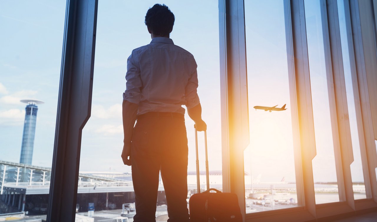 Person steht mit Handgepäck am Fenster eines Flughafens und blickt auf startendes Flugzeug bei Sonnenuntergang.