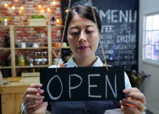 Eine Frau in einem Café oder kleinen Geschäft hängt ein Schild mit der Aufschrift „OPEN“ an die Tür. Im Hintergrund ist ein gemütlich eingerichteter Verkaufsraum mit Regalen und einer Menütafel zu sehen.