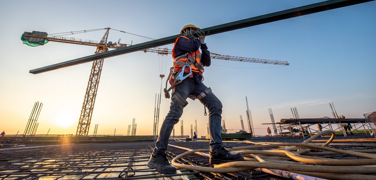 Das Bild zeigt eine Baustelle während des Sonnenuntergangs. Im Vordergrund trägt ein Bauarbeiter in Schutzkleidung, bestehend aus Helm, Sicherheitsweste und Klettergurt, einen langen Stahlträger auf der Schulter. Er steht auf einem Gitter aus Stahlarmierungen, das Teil des Baufundaments ist. Im Hintergrund sind mehrere Baukräne und weitere Arbeiter zu sehen. Die Szene wird von der warmen, goldenen Abendsonne beleuchtet, die lange Schatten wirft.