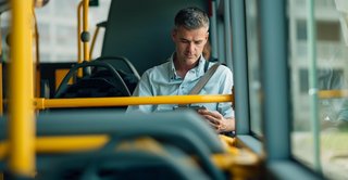 Ein Mann mittleren Alters sitzt in einem fast leeren Linienbus und schaut auf sein Smartphone. Er trägt ein helles Hemd und eine Umhängetasche, während Sonnenlicht durch die Fenster fällt. Die Atmosphäre wirkt ruhig und konzentriert.