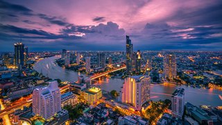 Panoramablick über eine moderne Großstadt bei Sonnenuntergang. Entlang eines breiten Flusses stehen zahlreiche beleuchtete Hochhäuser und Brücken, deren Lichter sich im Wasser spiegeln. Der Himmel zeigt ein intensives Farbspiel aus Violett, Rosa und Blau mit dramatischen Wolkenformationen.
