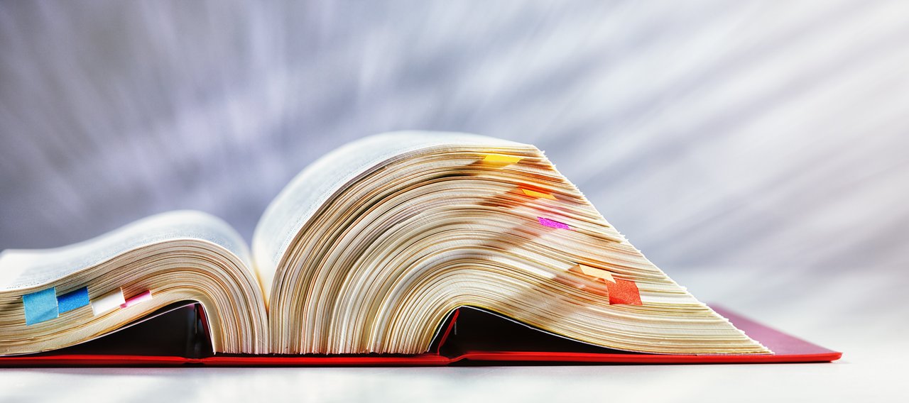 Ein dickes, aufgeschlagenes Gesetzbuch liegt auf einer hellen Fläche. Die Seiten sind mit zahlreichen farbigen Klebezetteln markiert, die aus dem Buch herausragen. Der Hintergrund ist weich verschwommen und strahlt in hellen Tönen.