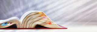 Ein dickes, aufgeschlagenes Gesetzbuch liegt auf einer hellen Fläche. Die Seiten sind mit zahlreichen farbigen Klebezetteln markiert, die aus dem Buch herausragen. Der Hintergrund ist weich verschwommen und strahlt in hellen Tönen.