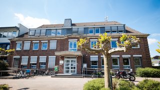 Das Bild zeigt das Rathaus von Rommerskirchen, ein zweistöckiges Backsteingebäude mit Walmdach und weißen Fensterrahmen. Vor dem Eingang stehen Fahrräder und ein Motorrad, daneben sind kleine Bäume mit frischem Austrieb und Büsche zu sehen. Der Himmel ist blau mit wenigen Wolken, was eine freundliche, sonnige Atmosphäre vermittelt.