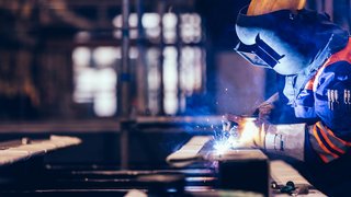 „Person führt Schweißarbeiten in Industriehalle durch. Sie trägt Schutzmaske, gelben Helm, feuerfeste Handschuhe und Arbeitsjacke mit orangefarbenen Streifen. Funken fliegen, bläulicher Rauch steigt auf. Unscharfer Hintergrund deutet industrielle Umgebung an.