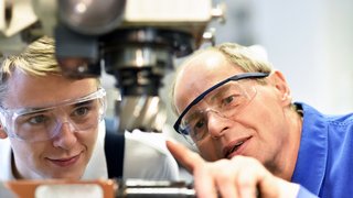 Zwei Personen mit Schutzbrillen arbeiten gemeinsam an einer Maschine; eine Person zeigt mit der Hand auf einen Bereich der Anlage.