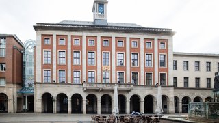 Frontalansicht des Rathauses von Neuss auf dem Marktplatz. Das mehrstöckige Gebäude besitzt eine symmetrische Fassade aus roten und hellen Steinflächen mit hohen rechteckigen Fenstern. Im Erdgeschoss verläuft eine Arkade mit mehreren Rundbögen. Über dem mittleren Teil befindet sich ein Balkon sowie ein Wappen. Auf dem Dach ragt ein Uhrturm mit blauem Zifferblatt empor. Vor dem Gebäude stehen Tische und Stühle eines Außenbereichs sowie ein Brunnen im rechten Vordergrund.
