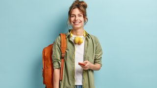 Eine junge Frau steht vor einem hellblauen Hintergrund. Sie trägt ein grünes Hemd über einem weißen Oberteil, blaue Jeans und hat einen orangefarbenen Rucksack über einer Schulter sowie gelbe Kopfhörer um den Hals. Sie hält ein Smartphone in einer Hand, mit dem Kopfhörerkabel verbunden.