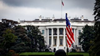 „Das Weiße Haus in Washington, D.C. bei bewölktem Himmel. Im Vordergrund weht eine amerikanische Flagge an einem Mast. Am unteren Bildrand ist der angeschnittene Kopf einer Person zu sehen. Die Szene wirkt ernst und leicht düster.