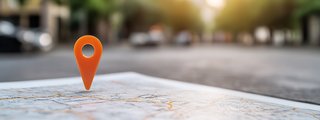 Nahaufnahme einer Landkarte mit einem orangefarbenen Standort-Pin darauf; im unscharfen Hintergrund eine städtische Straße mit Bäumen und Gebäuden bei Sonnenuntergang.