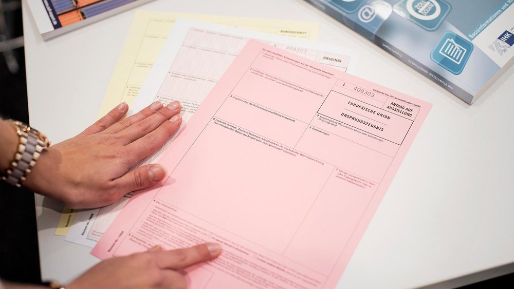 Hände einer Person, die ein rosa Formular mit der Aufschrift „Antrag auf Ausstellung Europäische Union Ursprungszeugnis“ auf einem Tisch hält, daneben weitere Formulare und Broschüren.