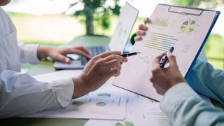Zwei Personen sitzen an einem Tisch und analysieren Geschäftsdaten. Eine Person hält ein Klemmbrett mit Diagrammen und Grafiken, während beide mit Stiften auf die Daten zeigen. Auf dem Tisch liegen weitere Auswertungen und im Hintergrund ist ein Laptop zu sehen.