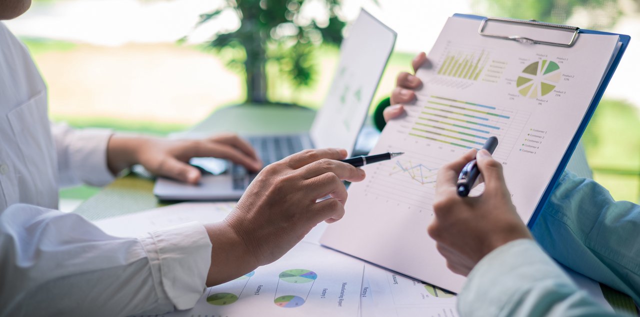 Zwei Personen sitzen an einem Tisch und analysieren Geschäftsdaten. Eine Person hält ein Klemmbrett mit Diagrammen und Grafiken, während beide mit Stiften auf die Daten zeigen. Auf dem Tisch liegen weitere Auswertungen und im Hintergrund ist ein Laptop zu sehen.