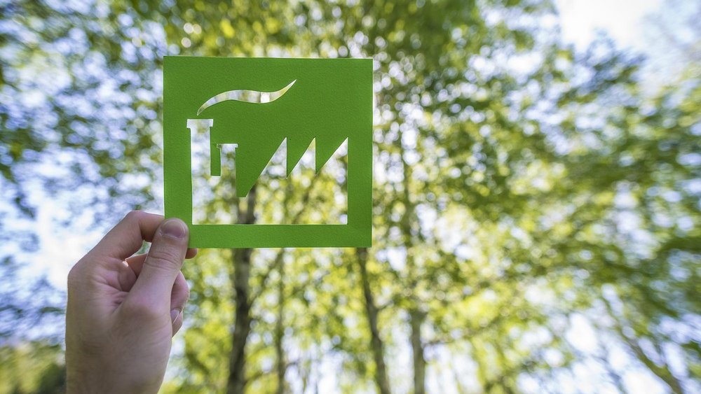 Eine Person hält eine grüne, rechteckige Papierschablone mit einer ausgestanzten Fabriksilhouette vor einen Hintergrund aus unscharfen, leuchtend grünen Bäumen. Der Fabrikumriss zeigt Schornsteine und ein gezacktes Dach. Der Rauch aus dem Schornstein ist ebenfalls ausgeschnitten, sodass der natürliche Hintergrund durchscheint. Die Szene vermittelt eine Botschaft zur Nachhaltigkeit und umweltfreundlichen Industrie.