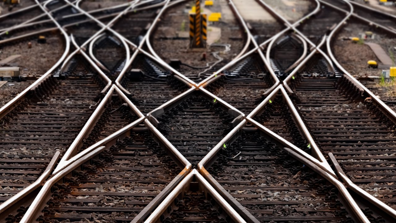 Mehrere Eisenbahngleise mit komplexem Weichensystem, das sich kreuzt und verzweigt. Die Schienen liegen auf dunklem, steinigem Untergrund und führen in verschiedene Richtungen, symbolisieren Verbindungen und Entscheidungswege.